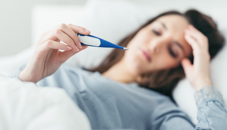 Žena koja ima prehladu leži u krevetu i gleda u termometar